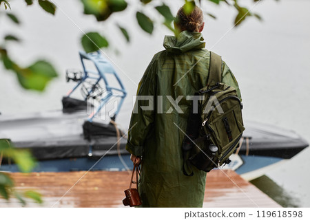 Back view of male tourist wearing green raincoat looking out at water while standing on pier in nature shot through leaves 119618598
