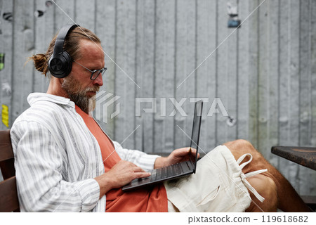 Minimal side view portrait of contemporary bearded man using laptop and wearing big headphones while working online or using internet sitting outdoors 119618682