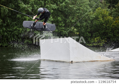 Wide angle action shot of sportsman enjoying wakeboarding on lake and doing high jumps on ramp with water splashes copy space 119618754