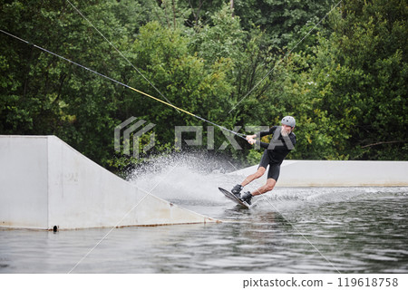Wide angle action shot of bearded active man enjoying wakeboarding and jumping onto ramp with water splashes copy space 119618758
