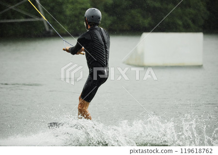 Back view of sportive adult man wearing wetsuit and practicing wakeboarding on lake waters moving towards ramps for action tricks copy space 119618762
