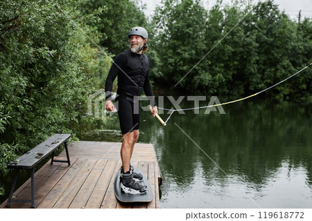 Candid full length shot of active senior man practicing wakeboarding on wooden pier outdoors and smiling 119618772