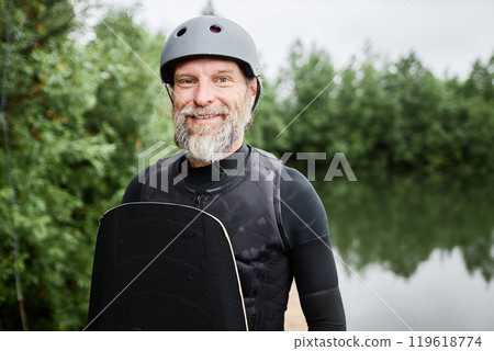 Waist up portrait of bearded senior man wearing wakeboarding gear and helmet smiling at camera outdoors standing by scenic forest lake copy space 119618774