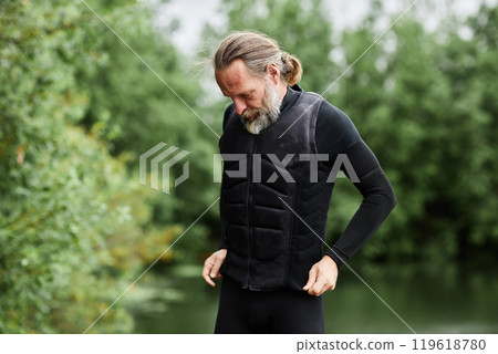 Waist up portrait of bearded active senior man putting on wetsuit and diving gear standing by scenic forest lake copy space 119618780