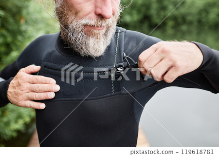 Close up of bearded mature man putting on wetsuit by lake and closing velcro fastenings getting ready for diving Close up of bearded mature man putting on wetsuit by lake and closing velcro fastenings getting ready for diving 119618781