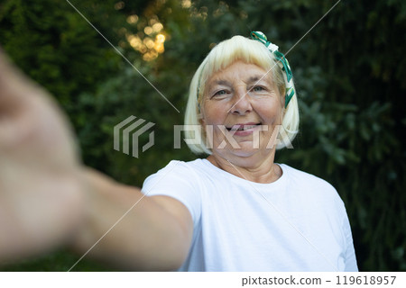 Portrait of stylish old lady in white t-shirt looking at camera and smiling, make selfie by camera in nature Portrait of stylish old lady in white t-shirt looking at camera and smiling, make selfie by camera in nature 119618957