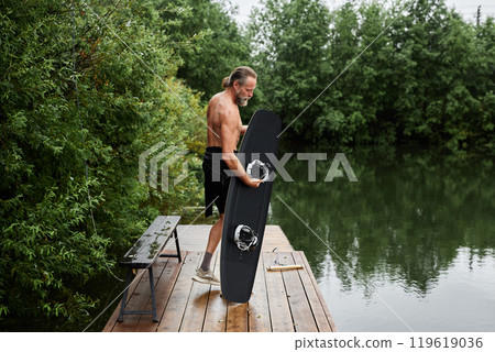 Full length shot of shirtless active senior man holding wakeboarding board standing on wooden pier by scenic forest lake copy space Full length shot of shirtless active senior man holding wakeboarding board standing on wooden pier by scenic forest lake copy space 119619036