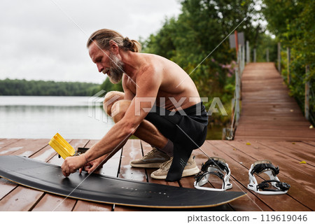 Full length side view portrait of active mature man cleaning and polishing wakeboarding equipment on wooden pier by lake copy space Full length side view portrait of active mature man cleaning and polishing wakeboarding equipment on wooden pier by lake copy space 119619046