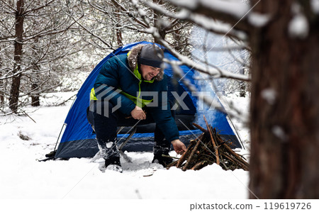 A happy tourist smiles by a fire in a snowy forest, enjoying the peace and beauty of nature. A man sits next to a tent in a winter forest and makes a fire. The man made a fire to warm up in winter. 119619726