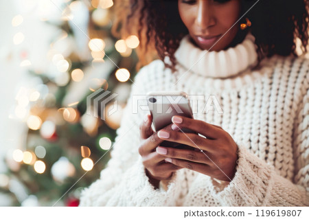 Black woman in cozy sweater texting on smartphone against festive background. Christmas, New Year concept 119619807
