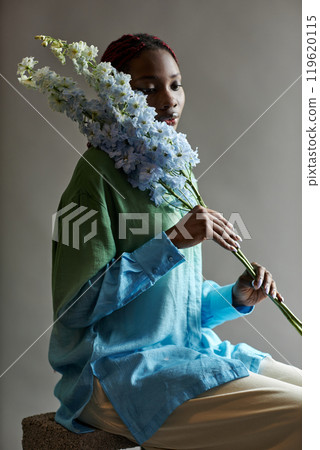 Delicate portrait of young Black woman posing with exotic blue flower accentuating femininity and grace in low lighting 119620115