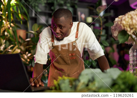 Portrait of young African American man using laptop in flower shop and managing flower delivery orders with neon light accent copy space 119620261