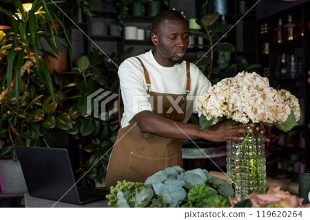 Waist up portrait of young African American man as male florist putting flowers in vase while opening elegant flower shop in morning copy space 119620264