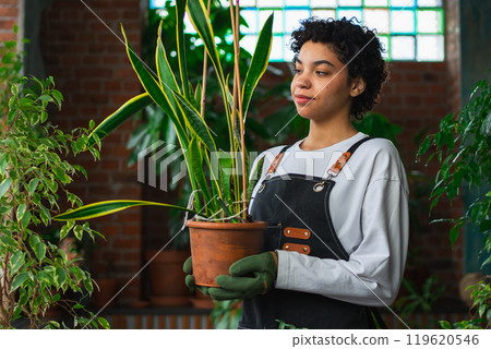 Running of own business. African woman florist holding plant in pot wearing apron in botanical store. Happy small business owner working at flower shop smiling surrounded by plants. Small business 119620546