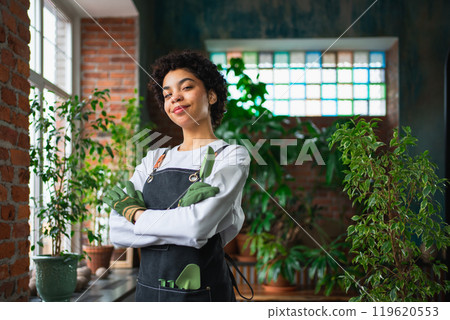 Running of own business. African woman florist wearing apron in botanical store with green plants. Happy small business owner working at flower shop smiling surrounded by plants Small business Running of own business. African woman florist wearing apron in botanical store with green plants. Happy small business owner working at flower shop smiling surrounded by plants Small business 119620553