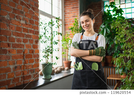 Running of own business. Young woman florist wearing apron in botanical store with green plants. Happy small business owner working at flower shop standing surrounded by plants. Small business 119620554