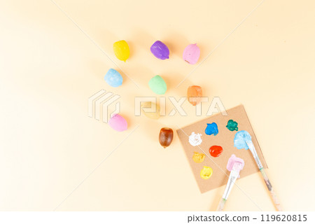 few painted acorns with emoji faces next to unpainted acorns, paint palette, and brushes on peach background. Flat lay autumn craft idea for cards, DIY nature craft project, autumn decoration, 119620815