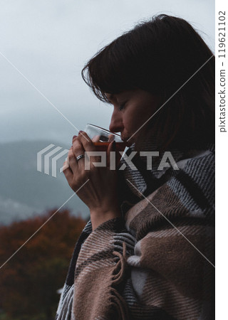 Girl drinking hot tea in checkered plaid on foggy mountains landscape. Traveler resting in autumn mountains. Woman in blanket dreaming in countryside. Travel in Carpathian mountains. Autumn mood. Girl drinking hot tea in checkered plaid on foggy mountains landscape. Traveler resting in autumn mountains. Woman in blanket dreaming in countryside. Travel in Carpathian mountains. Autumn mood. 119621102
