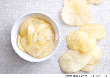 Salted potato chips in a white bowl on linen fabric. Fresh and ready to eat crisps, slim slices of deep fried potato, made from tubers of the root vegetable Solanum tuberosum. Close-up, from above. Salted potato chips in a white bowl on linen fabric. Fresh and ready to eat crisps, slim slices of deep fried potato, made from tubers of the root vegetable Solanum tuberosum. Close-up, from above. 119621161