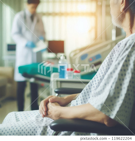 Senior patient in a hospital wheelchair with hands clasped, doctor blurred in the background with medical equipment and bed visible 119622204