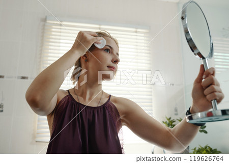 Young Woman Cleansing Face with Mirror in Modern Bathroom Young Woman Cleansing Face with Mirror in Modern Bathroom 119626764