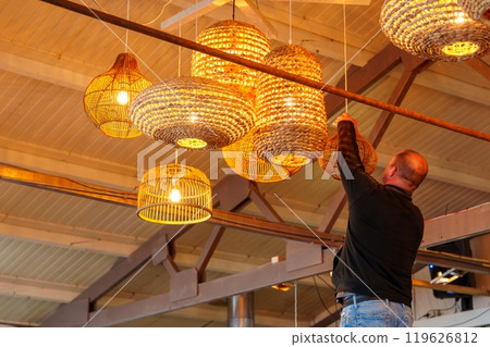 Male worker skillfully installing elegant wicker lamps in a cozy beachside restaurant setting. Male worker skillfully installing elegant wicker lamps in a cozy beachside restaurant setting. 119626812