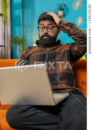 Upset Indian man sitting on home sofa using laptop having anxiety stress loss bad fail news virus Upset Indian man sitting on home sofa using laptop having anxiety stress loss bad fail news virus 119629280