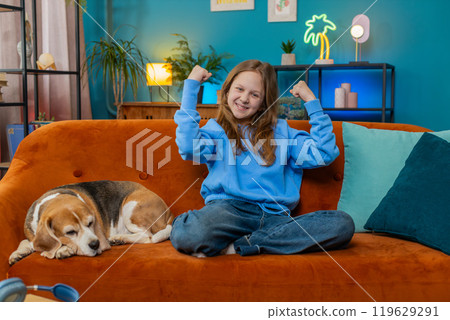 Cheerful young child girl celebrating victory winning the lottery at home beside beagle dog on sofa Cheerful young child girl celebrating victory winning the lottery at home beside beagle dog on sofa 119629291