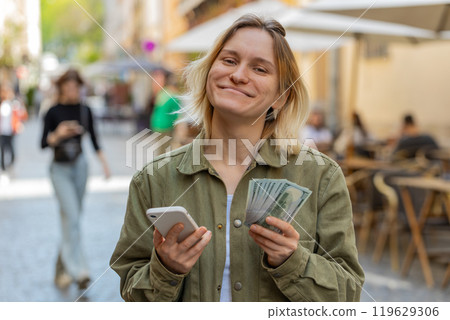 Woman counting money dollar cash using smartphone calculator app satisfied of salary on city street 119629306