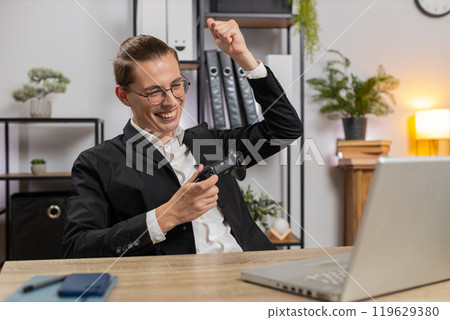 Excited young businessman playing video game on laptop with joystick controller at home office desk 119629380