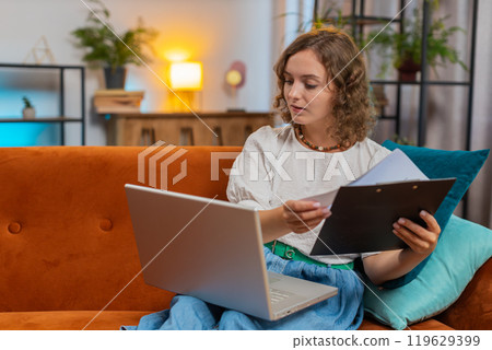 Woman accountant using laptop working with financial graph documents, reports of home income on sofa 119629399