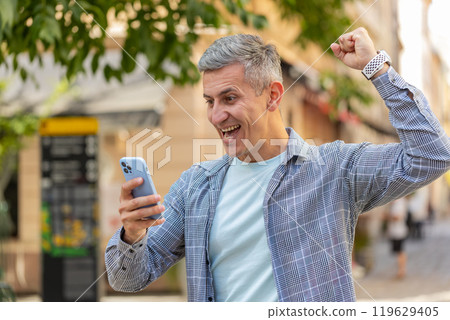 Happy Caucasian mature man using smartphone celebrating win good news, lottery on city town street Happy Caucasian mature man using smartphone celebrating win good news, lottery on city town street 119629405