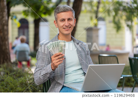 Happy rich man using laptop winner victory holding cash money in dollar banknotes sitting on chair Happy rich man using laptop winner victory holding cash money in dollar banknotes sitting on chair 119629408