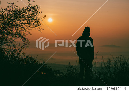A solitary figure stands softly silhouetted against the warm, glowing hues of an autumn sunrise. Mist rises from the valley as nature awakens to a new day filled with tranquility. A solitary figure stands softly silhouetted against the warm, glowing hues of an autumn sunrise. Mist rises from the valley as nature awakens to a new day filled with tranquility. 119630444