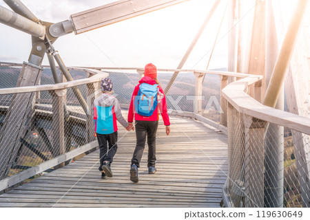 Two children walk hand in hand along the wooden pathway of the Sky Walk Tower in Swieradow Zdroj, enjoying a scenic view of the Jizera Mountains on a sunny day. 119630649