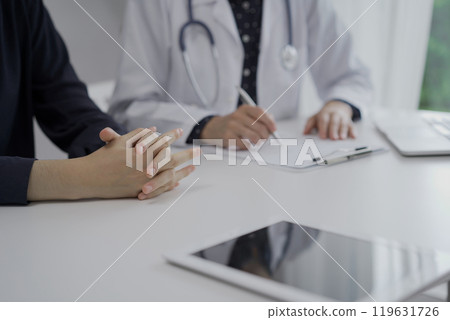 Doctor and a patient are sitting at the white table in clinic. The female physician is using clipboard for making notes during a consultation, close up of woman hands. Medicine concept 119631726