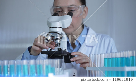 A female scientist, wearing a lab coat and safety glasses, is adjusting a binocular microscope surrounded by test tubes in laboratory setting. Medicine and science A female scientist, wearing a lab coat and safety glasses, is adjusting a binocular microscope surrounded by test tubes in laboratory setting. Medicine and science 119631919