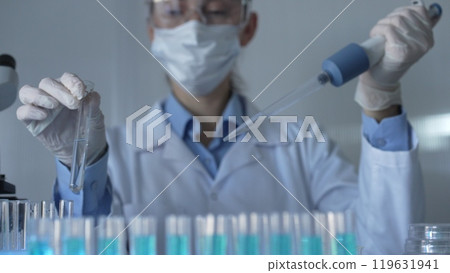 Scientist woman, wearing a lab coat, mask, blue gloves and safety glasses, is using a micropipette to drop a liquid into a test tubes. Medicine and science Scientist woman, wearing a lab coat, mask, blue gloves and safety glasses, is using a micropipette to drop a liquid into a test tubes. Medicine and science 119631941