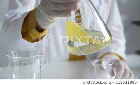 Unknown scientist wearing white protective gloves is carefully shaking up a yellow chemical solution inside an Erlenmeyer flask in a laboratory, close up. Science and medicine 119631980