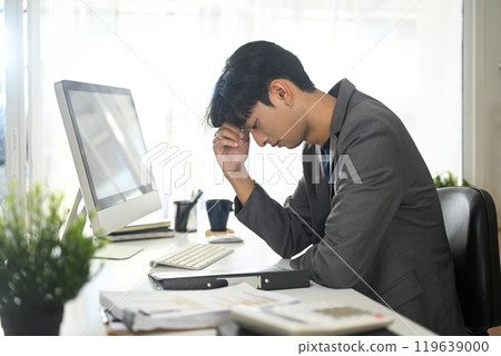 Frustrated young businessman in a business suit holding his head in hands sitting at desk 119639000
