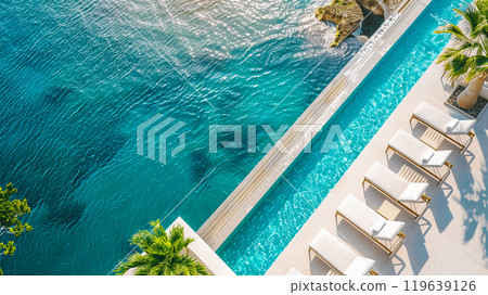 Aerial view of a luxurious infinity pool overlooking the ocean with several lounge chairs and tropical palm trees on a sunny day. 119639126