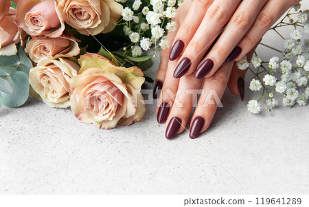 Elegant hands with dark manicure resting beside blush roses and white blooms on a light surface 119641289