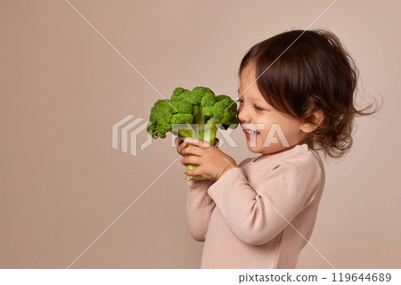 smiling little child girl holding fresh green raw broccoli on beige background. healthy baby food. kid loves vegetables smiling little child girl holding fresh green raw broccoli on beige background. healthy baby food. kid loves vegetables 119644689