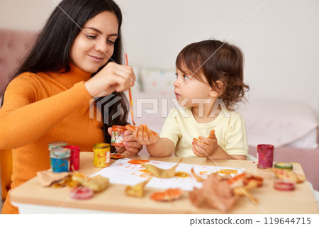 Cute little girl and mom spending good time painting hands and autumn yellow leaves together Cute little girl and mom spending good time painting hands and autumn yellow leaves together 119644715