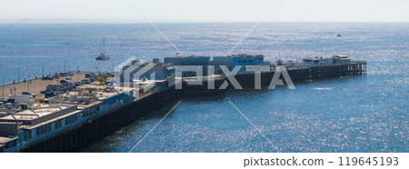 The Santa Cruz Wharf stretches into the Pacific Ocean, with buildings and parked cars along its length. Boats are visible in the distance under sunlight. 119645193