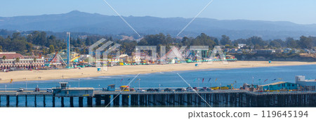 Aerial view of Santa Cruz, California, featuring the iconic beach boardwalk with colorful rides, the wharf extending into the ocean, and distant mountains. 119645194
