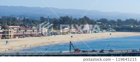 The image captures the Santa Cruz Beach Boardwalk with colorful rides, a large roller coaster, and a pier. The Pacific Ocean and green hills are visible. 119645215