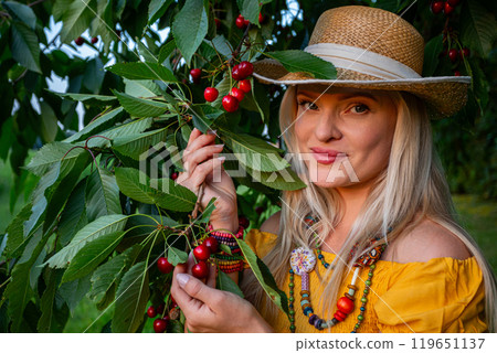 Smiling Woman Holding Ripe Red Cherries Beneath a Cherry Tree on a Warm Spring Day with Low-Hanging Branches 119651137