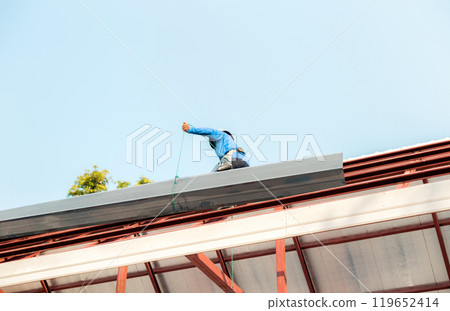 Roofer working in special protective work wear gloves, using air or pneumatic nail gun installing asphalt shingle on top of the new roof, Concept of residential building under construction. 119652414