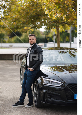 Handsome bearded man stand by his luxury car near the home. Stylish male in leather jacket  119653326